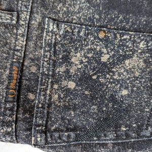 May include: Black denim jeans with a white splatter paint design. The jeans have a back pocket with a small brown leather patch that says "Wrangler" in white stitching.