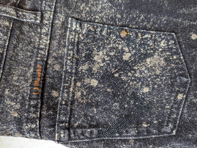 May include: Black denim jeans with a white splatter paint design. The jeans have a back pocket with a small brown leather patch that says "Wrangler" in white stitching.