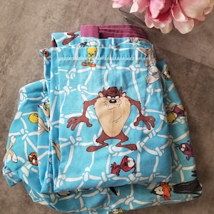 Op de afbeelding: Een blauwe en witte stof met een herhalend patroon van een cartoonfiguur, Tweety Bird, Taz en Bugs Bunny. De stof is dubbelgevouwen en heeft een paarse en witte rand.
