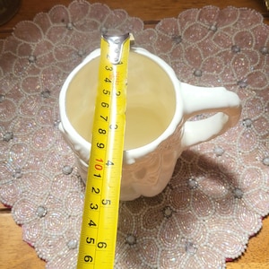 May include: A white ceramic mug with a handle, measured with a yellow tape measure. The mug is approximately 4 inches (10 cm) tall. The mug sits on a pink beaded placemat.