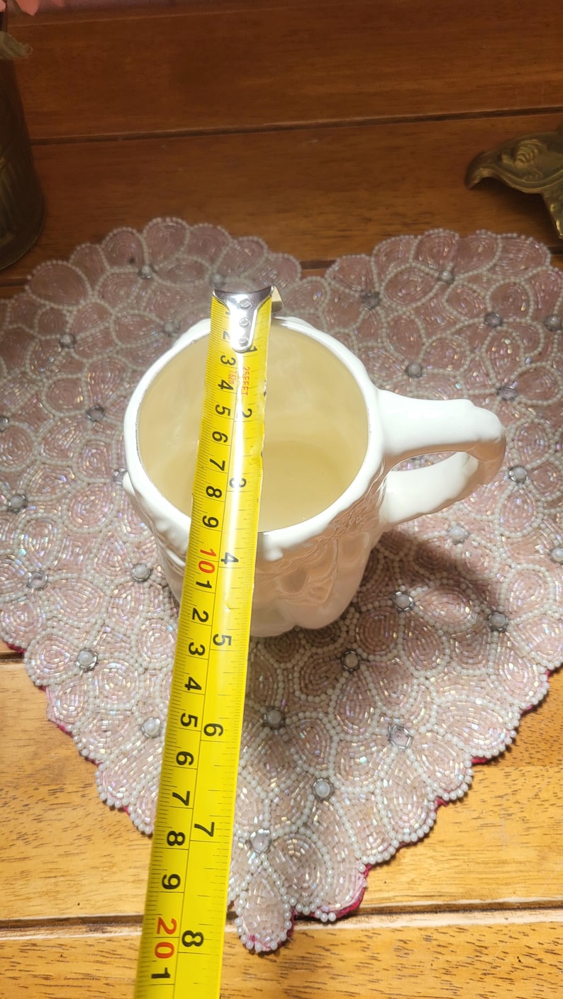 May include: A white ceramic mug with a handle, measured with a yellow tape measure. The mug is approximately 4 inches (10 cm) tall. The mug sits on a pink beaded placemat.