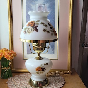 以下が含まれることがあります： オレンジ、茶色、クリーム色のバラの花柄が施された、ヴィンテージの白いガラスランプ。 真鍮製のベースとアクセント、お揃いのシェードが付いています。 ランプは木製の表面に置かれ、その下に装飾的なドイリーがあります。