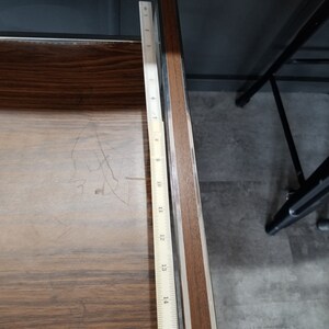 May include: A close-up of a wooden surface with a metal ruler measuring 16 inches. The ruler is placed along the edge of the surface, which is brown with a wood grain pattern.