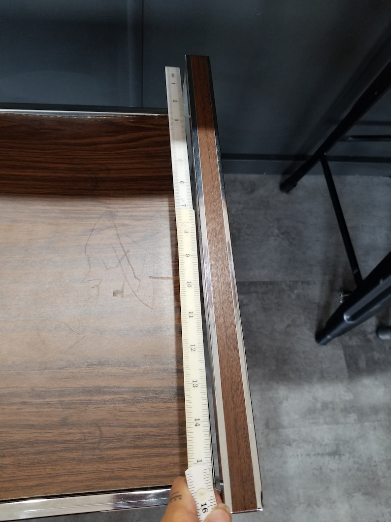 May include: A close-up of a wooden surface with a metal ruler measuring 16 inches. The ruler is placed along the edge of the surface, which is brown with a wood grain pattern.