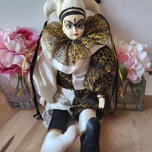 May include: A porcelain jester doll with a white and black costume, gold trim, and a black butterfly mask. The doll is sitting on a wooden surface with flowers in vases behind it.