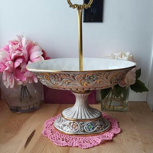 Op de afbeelding: Een witte keramische serveerschaal met gouden accenten en bloemmotieven. De schaal heeft een voet en een afneembare gouden handgreep. De schaal staat op een roze gehaakt onderzetter.
