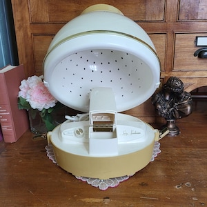 May include: Vintage Lady Sunbeam hair dryer in cream and gold. The dryer has a domed hood with ventilation holes, a control panel, and a base. The dryer is sitting on a wooden surface.