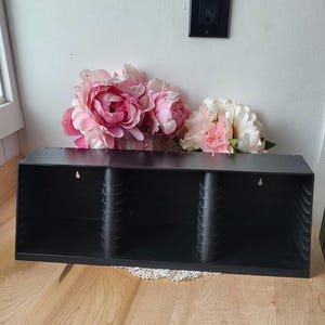 May include: Black wall-mounted CD storage unit with three compartments. The unit is empty and has a hole at the top for hanging.