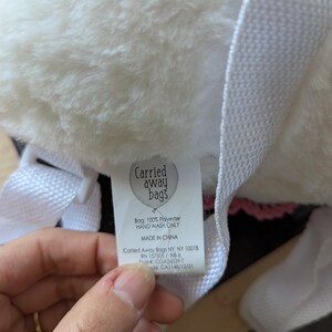 May include: White faux fur backpack with a pink trim and a white tag that reads "Carried away bags" with the text "Bag: 100% Polyester Hand Wash Only Made in China Carried Away Bags NY, NY 10018 RN 157505 / NB-6 Style#: CGA26029-1 Made: CA1148/12/21"