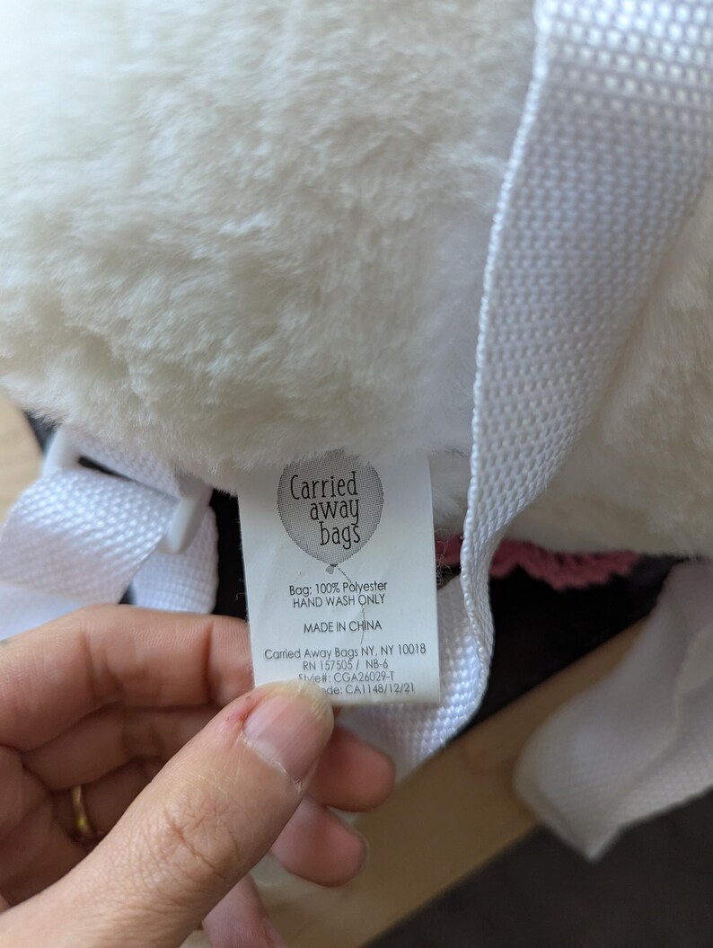May include: White faux fur backpack with a pink trim and a white tag that reads "Carried away bags" with the text "Bag: 100% Polyester Hand Wash Only Made in China Carried Away Bags NY, NY 10018 RN 157505 / NB-6 Style#: CGA26029-1 Made: CA1148/12/21"