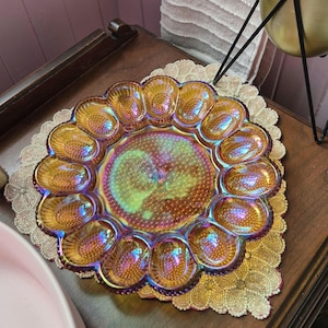 Puede incluir: Un plato para huevos redondo de vidrio de carnaval iridiscente color caléndula, con un patrón central de puntos rodeado por doce hendiduras ovaladas individuales a lo largo del borde festoneado, con un acabado metálico multicolor.