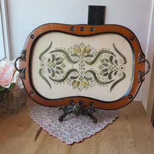 Puede incluir: Una bandeja ovalada para servir con marco de madera y asas decorativas de metal. El interior de la bandeja presenta una superficie crema con un diseño floral en verde y dorado. La bandeja se exhibe sobre un soporte decorativo de metal.