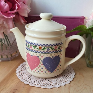 May include: A cream-coloured teapot with a floral design featuring pink and purple hearts. The teapot has a blue and white checked band around the middle and a floral design with small hearts.
