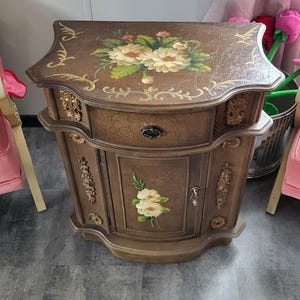 May include: A brown, ornate nightstand with a floral design on the top and front. The nightstand has a drawer and a cabinet door, with gold-colored accents and hardware. The top is a lighter brown with a crackled texture.
