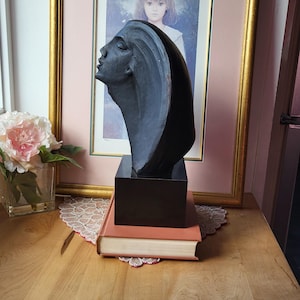 Op de afbeelding: Een donkergrijze sculptuur van het profiel van een vrouw, gemonteerd op een zwarte basis, rust op een stapel boeken. De sculptuur staat tegenover een ingelijst schilderij en een vaas met roze bloemen, wat een klassieke esthetiek creëert.