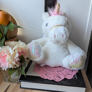 May include: White plush unicorn toy with pink and gold accents sitting on a pink doily on top of a black book.