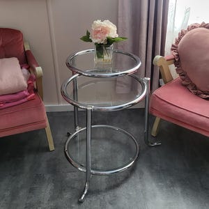 May include: A chrome and glass side table with three circular tiers. A vase of pink flowers sits on the top tier. The table is in a room with pink chairs and a framed picture on the wall.