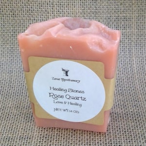 May include: A pink, rectangular bar of soap with a rounded top and a small rose quartz stone embedded on top. A label wraps around the soap, with the text "Healing Stones Rose Quartz Love & Healing" and "NET WT (4 OZ)".