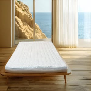 Puede incluir: Un colchón blanco con un diseño acolchado sobre una estructura de cama de madera. La cama está en una habitación con una gran ventana con vistas al mar. La habitación tiene suelos de madera y cortinas blancas.