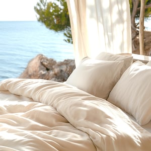 May include: White bedding with a white duvet cover and two white pillows on a bed with a view of the ocean through a sheer curtain.