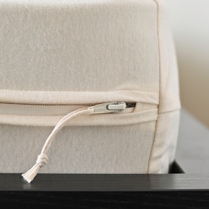 May include: Close-up of a beige fabric cushion with a zipper closure. The zipper is silver and has a fabric pull. The cushion appears to be made of a textured, natural-colored material.