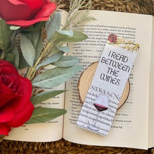 Może przedstawiać: Biała zakładka z kieliszkiem czerwonego wina i tekstem "I read between the wines". Zakładka leży na drewnianym podkładce i książce z czerwonymi różami.