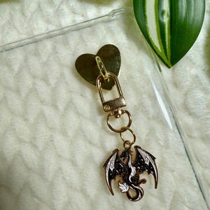 May include: A gold-coloured keychain featuring a heart-shaped clasp and a dragon charm. The dragon charm is black and white with detailed wings and body. The keychain is attached to a clear phone case.