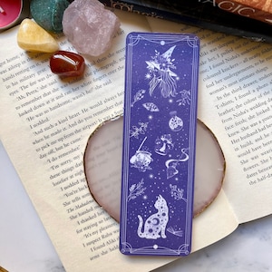 May include: Purple bookmark with a white illustration of a witch, a cat, and a broom. The bookmark has a white border and a white background with a starry pattern. The text "PRACTICAL MAGIC" is printed on the top of the bookmark.
