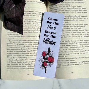 May include: A white bookmark with a black snake wrapped around red roses. The bookmark has the text "Came for the Hero Stayed for the Villain" in black text.