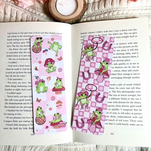 May include: Two pink and white checkered bookmarks with a frog wearing a cowboy hat and holding a lasso. The frog is on a mushroom with a pink and white checkered background. The other bookmark has a frog wearing a pink hat and holding a cup of tea. The frog is on a disco ball with a pink and white checkered background. Both bookmarks have a pink and white checkered border.