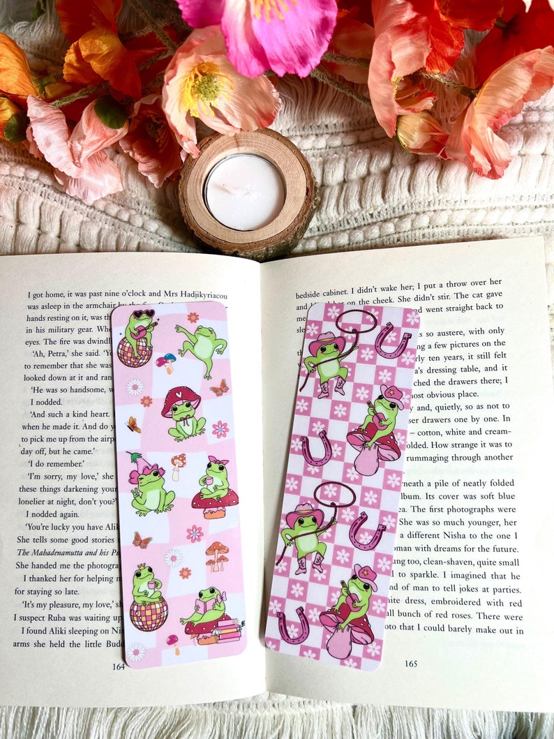 May include: Two pink and white checkered bookmarks with a frog wearing a cowboy hat and holding a lasso. The frog is on a mushroom with a pink and white checkered background. The other bookmark has a frog wearing a pink hat and holding a cup of tea. The frog is on a disco ball with a pink and white checkered background. Both bookmarks have a pink and white checkered border.