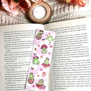 May include: A close-up of a bookmark with a pink and white checkered background. The bookmark features a cartoon frog wearing a hat and sunglasses, holding a guitar and sitting on a mushroom. The text "Nisha" is visible at the bottom of the bookmark.