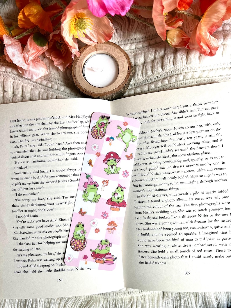 May include: A close-up of a bookmark with a pink and white checkered background. The bookmark features a cartoon frog wearing a hat and sunglasses, holding a guitar and sitting on a mushroom. The text "Nisha" is visible at the bottom of the bookmark.