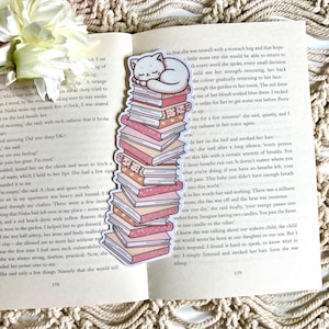 Peut inclure: Un chat blanc aux yeux fermés dort sur une pile de livres roses et blancs. Le chat est enroulé sur le livre du haut, la tête reposant sur ses pattes. Les livres sont empilés en une tour, le livre du haut étant le plus grand. Les livres sont de tailles et de formes différentes, et ils sont tous décorés de fleurs roses et blanches. Le chat est un autocollant, et les livres sont un autocollant.