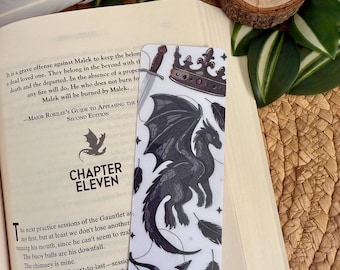 May include: A rectangular bookmark with a black dragon illustration, a crown, sword, bow and arrow, and feathers on a white background. The bookmark is placed on an open book.