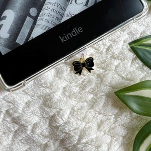 Pode incluir: Um marcador de página preto e dourado com um design de laço. O marcador de página está preso a um leitor de livros eletrônicos Kindle.