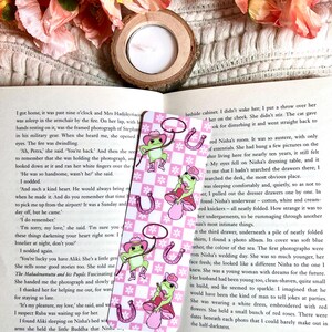 May include: A pink and white bookmark with a cartoon frog wearing a cowboy hat and holding a lasso. The frog is standing on a pink mushroom with white polka dots. The bookmark has a checkered pattern and a horseshoe design.