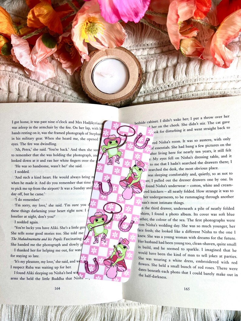 May include: A pink and white bookmark with a cartoon frog wearing a cowboy hat and holding a lasso. The frog is standing on a pink mushroom with white polka dots. The bookmark has a checkered pattern and a horseshoe design.