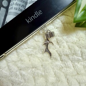 May include: A silver charm in the shape of an open book with a decorative flowing design. The charm rests on a black Kindle device, with the word "kindle" printed on it. The background is a soft, white, textured fabric.