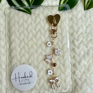 Könnte beinhalten: Transparente Handyhülle mit einem herzförmigen goldenen Verschluss und einer Charm-Kette. Die Kette hat weiße und goldene Charms, darunter Blumen, einen Schmetterling und eine Schleife. Ein weißer Aufkleber mit "Hooked on a Book" befindet sich auf der Hülle.