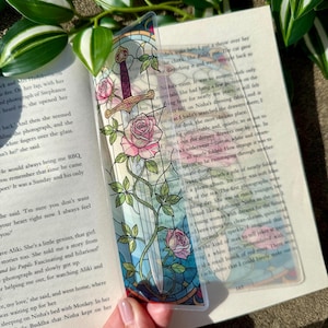 Op de afbeelding: Transparante acryl boekenlegger met een glas-in-lood ontwerp met een zwaard, roze rozen en groene wijnranken. De boekenlegger ligt in een open boek.
