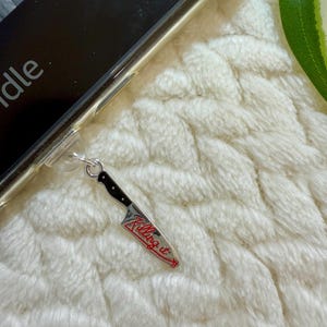 May include: A silver knife charm with a black handle and the words "Killing it" in red script. The charm is attached to a clear phone case, resting on a white, textured surface.