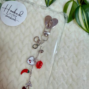 Pode incluir: Uma capa de telefone transparente com um pingente prateado com fecho em forma de coração e uma corrente de pingentes. Os pingentes incluem uma pimenta vermelha, uma rosa vermelha, um coração branco com cerejas vermelhas e um par de cerejas vermelhas.