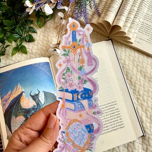 Puede incluir: Una pegatina vertical que muestra una espada dorada apuntando hacia arriba, apoyada sobre una pila de tres libros azules y rosas. Un huevo de dragón estampado descansa en la base, rodeado de flores rosas, ramas de lavanda y cintas moradas.