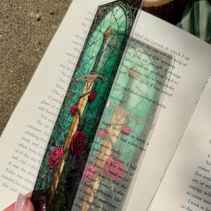 Op de afbeelding: Een decoratieve bladwijzer met een zwaardontwerp en rode rozen. De bladwijzer heeft een glas-in-lood effect met groene en gouden accenten. De bladwijzer ligt op een open boek.