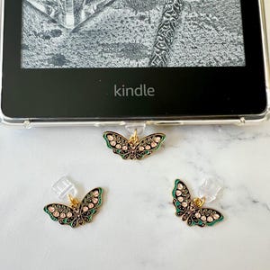 Op de afbeelding: Drie vlinderbedels met goudkleurig metaal en zwart email. De vlindervleugels hebben roze en groene bloemendetails. Het woord "kindle" is zichtbaar op het scherm van een e-reader.