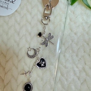 May include: A clear phone case with a silver heart-shaped charm and a chain of charms. The chain includes a dragonfly, a crescent moon, a star, a black heart, and an oval black stone charm. The background is a white, textured surface.
