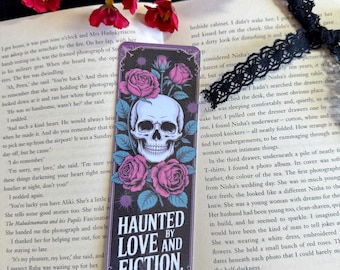 May include: Bookmark with a skull and pink roses design, with the text "HAUNTED BY LOVE AND FICTION." The bookmark is on a book page with red flowers in the background.