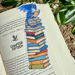 May include: A whimsical bookmark featuring a blue dragon perched atop a stack of colorful books, reading a smaller book. The bookmark is placed on an open book with the text "CHAPTER FIFTEEN". The dragon has detailed wings and is reading a book.