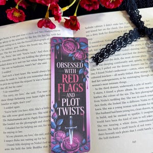 Peut inclure: Un marque-page avec le texte "Obsédé par les drapeaux rouges et les rebondissements" et une illustration de dague, entouré de roses roses et de feuilles. Le marque-page est posé sur un livre ouvert avec des fleurs rouges.